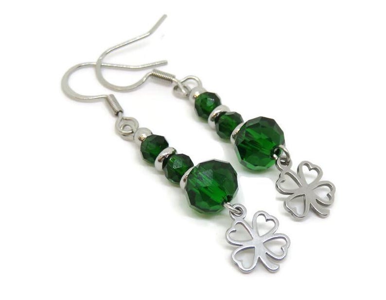 May include: A pair of dangle earrings featuring faceted green beads and silver-tone accents. Each earring has a silver-tone four-leaf clover charm. The earrings are displayed against a white background.
