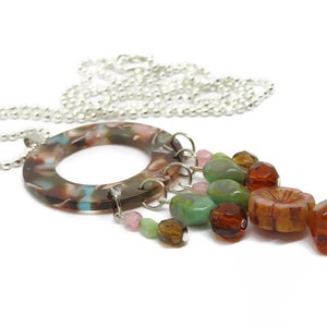 May include: A silver chain necklace with a brown, pink, and blue marbled circle pendant. The pendant has a cluster of colorful glass beads hanging from it, including green, brown, and pink.