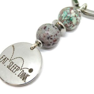 May include: A silver keychain with the text "EAT. SLEEP. DINK." on a round charm. The keychain features decorative beads in shades of gray, green, and silver. The design is simple and modern, suitable for everyday use.