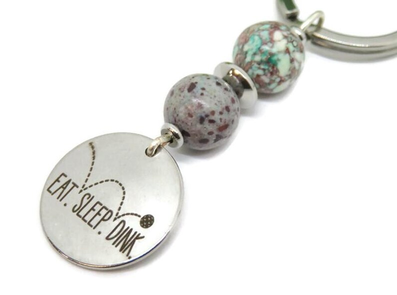 May include: A silver keychain with the text "EAT. SLEEP. DINK." on a round charm. The keychain features decorative beads in shades of gray, green, and silver. The design is simple and modern, suitable for everyday use.