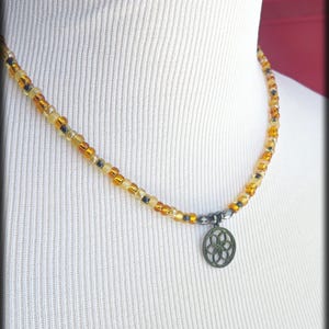 May include: A beaded necklace featuring a sequence of translucent amber, yellow, and dark grey beads. A circular dark metal charm with a geometric flower-like cutout pattern hangs from the center. The text MyCreativeSideJewels.etsy.com is visible.