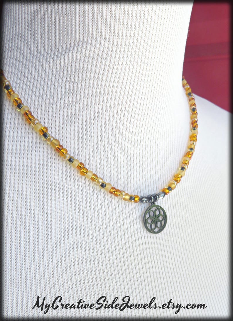 May include: A beaded necklace featuring a sequence of translucent amber, yellow, and dark grey beads. A circular dark metal charm with a geometric flower-like cutout pattern hangs from the center. The text MyCreativeSideJewels.etsy.com is visible.
