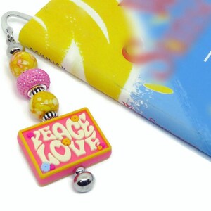 May include: A pink and yellow beaded bookmark with a pink charm that says "PEACE LOVE" in white letters. The charm has small yellow and pink flowers on it.