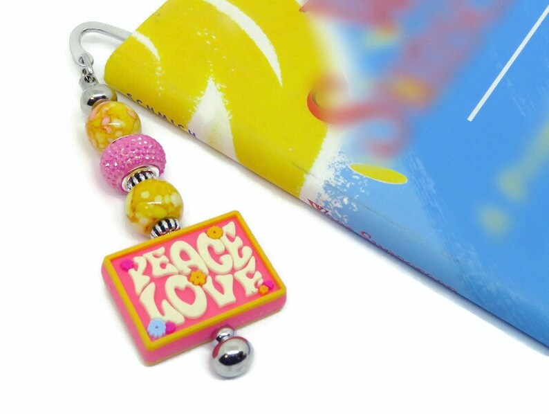 May include: A pink and yellow beaded bookmark with a pink charm that says "PEACE LOVE" in white letters. The charm has small yellow and pink flowers on it.
