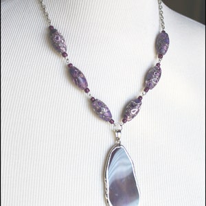 May include: A silver chain necklace with purple and white agate beads and a large purple agate pendant. The pendant is a natural stone with a unique pattern.