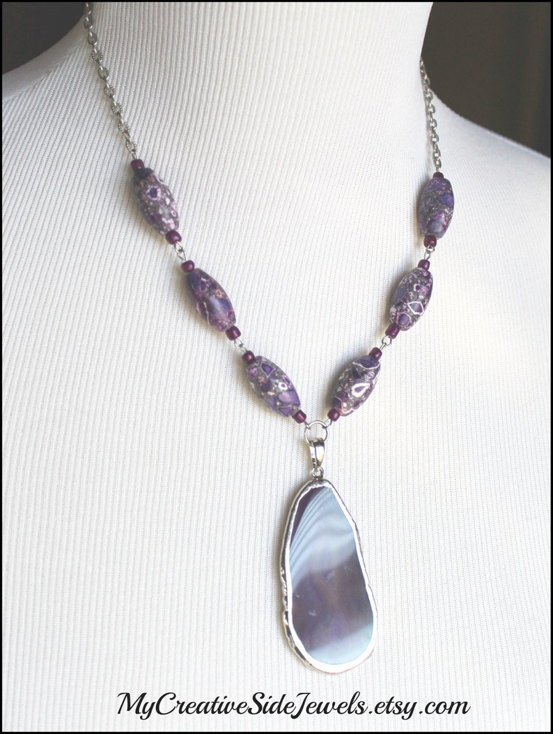 May include: A silver chain necklace with purple and white agate beads and a large purple agate pendant. The pendant is a natural stone with a unique pattern.