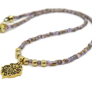 May include: A delicate necklace featuring a gold-colored pendant with an intricate black design. The necklace has a beaded chain in shades of lavender, gold, and brown. The clasp and connecting beads are gold.
