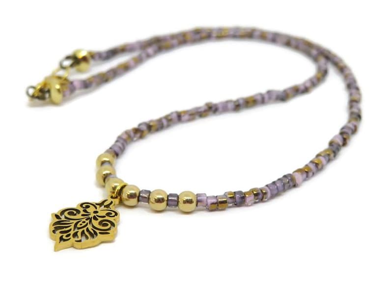 May include: A delicate necklace featuring a gold-colored pendant with an intricate black design. The necklace has a beaded chain in shades of lavender, gold, and brown. The clasp and connecting beads are gold.