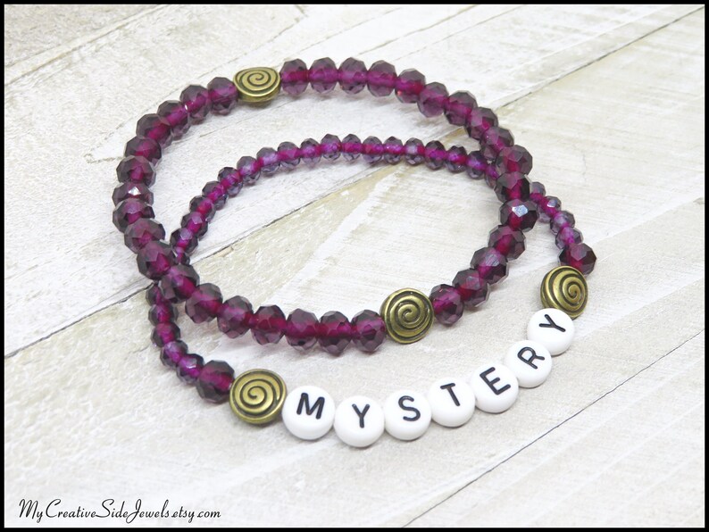 May include: Two purple crystal beaded bracelets with gold swirl charms and white letter beads spelling "MYSTERY".