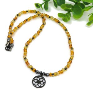 May include: A beaded necklace featuring a circular black pendant with a geometric flower-like cutout design. The strand consists of small, translucent amber, yellow, and brown glass beads interspersed with small, dark metallic gray accent beads.