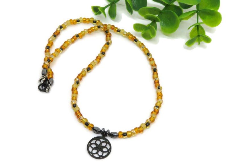 May include: A beaded necklace featuring a circular black pendant with a geometric flower-like cutout design. The strand consists of small, translucent amber, yellow, and brown glass beads interspersed with small, dark metallic gray accent beads.