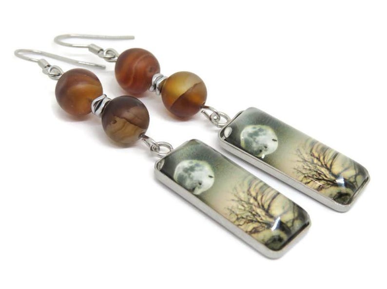 May include: A pair of dangle earrings with a silver tone finish. The earrings feature two brown agate beads and a rectangular charm with a moon and tree design.
