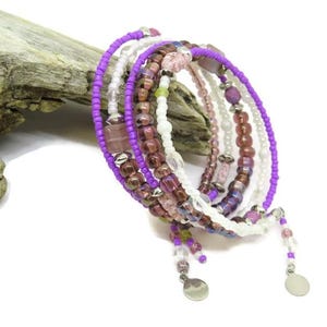 May include: A multi-strand wrap bracelet featuring a mix of purple, white, and burgundy beads. The bracelet includes small, round beads and larger, square and oval-shaped beads. Silver-colored charms dangle from the ends.