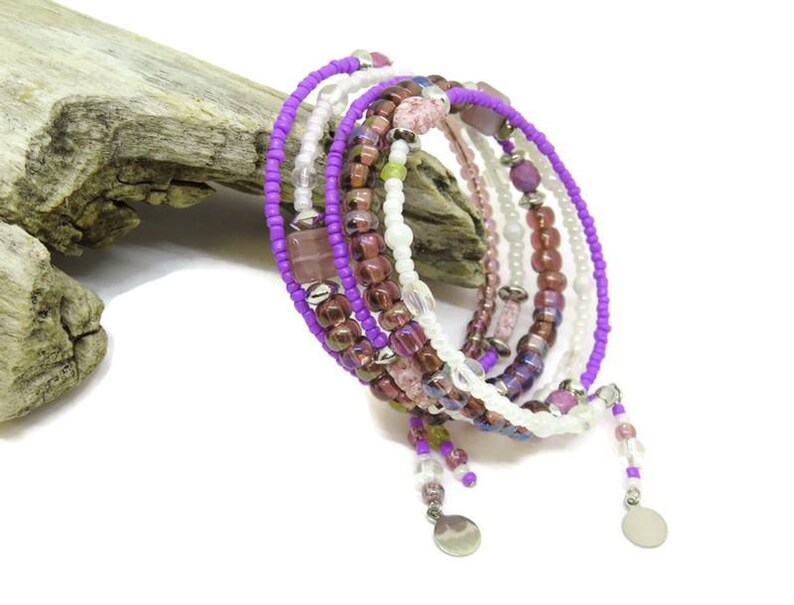 May include: A multi-strand wrap bracelet featuring a mix of purple, white, and burgundy beads. The bracelet includes small, round beads and larger, square and oval-shaped beads. Silver-colored charms dangle from the ends.