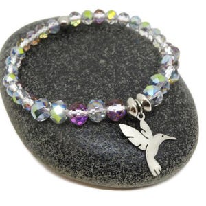 May include: A stretch bracelet featuring faceted, iridescent crystal beads in shades of clear, light purple, and yellow. The bracelet includes a silver-toned metal charm shaped like a hummingbird, attached to the bead strand with a silver-toned metal bead.