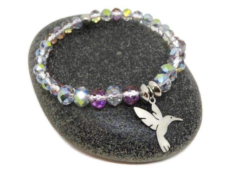 May include: A stretch bracelet featuring faceted, iridescent crystal beads in shades of clear, light purple, and yellow. The bracelet includes a silver-toned metal charm shaped like a hummingbird, attached to the bead strand with a silver-toned metal bead.