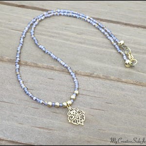May include: A delicate necklace featuring a double strand of small, light blue beads. A gold-colored pendant with an ornate floral design hangs from the center. The necklace is secured with a gold-colored clasp.