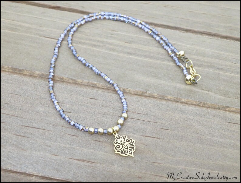 May include: A delicate necklace featuring a double strand of small, light blue beads. A gold-colored pendant with an ornate floral design hangs from the center. The necklace is secured with a gold-colored clasp.