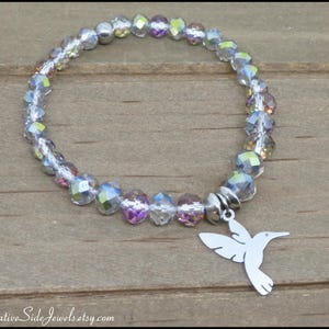 May include: A stretch bracelet featuring faceted, iridescent clear beads with a silver-toned hummingbird charm. The text MyCreativeSideJewels.etsy.com is visible at the bottom of the image. The beads reflect multiple colors including light pink and blue.