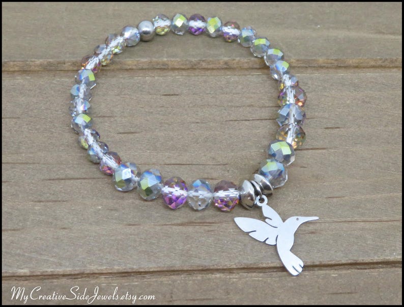 May include: A stretch bracelet featuring faceted, iridescent clear beads with a silver-toned hummingbird charm. The text MyCreativeSideJewels.etsy.com is visible at the bottom of the image. The beads reflect multiple colors including light pink and blue.