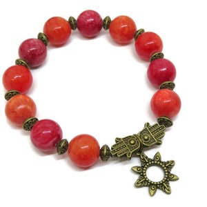 Pu&ograve; includere: Un braccialetto con perline rotonde rosse e arancioni, distanziatori decorativi color bronzo e un ciondolo a mano di Hamsa con un design a forma di sole. Il braccialetto &egrave; su sfondo bianco.