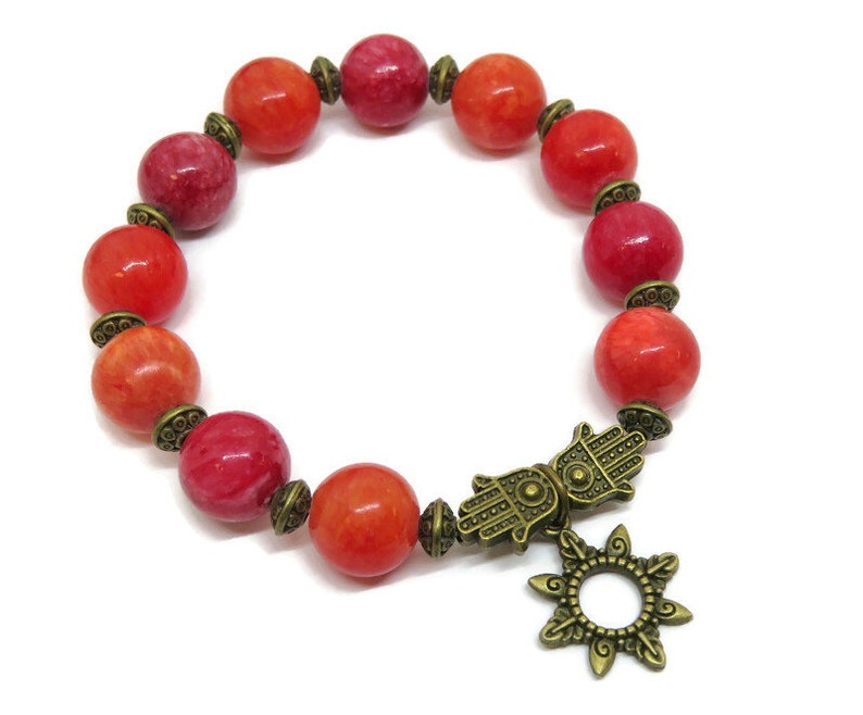 Pu&ograve; includere: Un braccialetto con perline rotonde rosse e arancioni, distanziatori decorativi color bronzo e un ciondolo a mano di Hamsa con un design a forma di sole. Il braccialetto &egrave; su sfondo bianco.