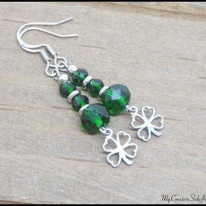 May include: A pair of dangle earrings featuring faceted emerald green beads and silver-tone clover charms. The earrings have silver-tone ear wires. The jewelry is displayed on a light brown wooden surface.