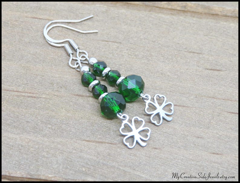 May include: A pair of dangle earrings featuring faceted emerald green beads and silver-tone clover charms. The earrings have silver-tone ear wires. The jewelry is displayed on a light brown wooden surface.