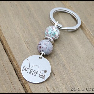 May include: A silver keychain with a round charm that reads "EAT. SLEEP. DINK." The keychain features two speckled beads in shades of purple and teal. The background is a wooden surface.