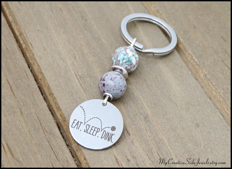 May include: A silver keychain with a round charm that reads "EAT. SLEEP. DINK." The keychain features two speckled beads in shades of purple and teal. The background is a wooden surface.