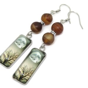 May include: Silver dangle earrings with brown agate beads and rectangular glass charms featuring a moon and tree illustration.
