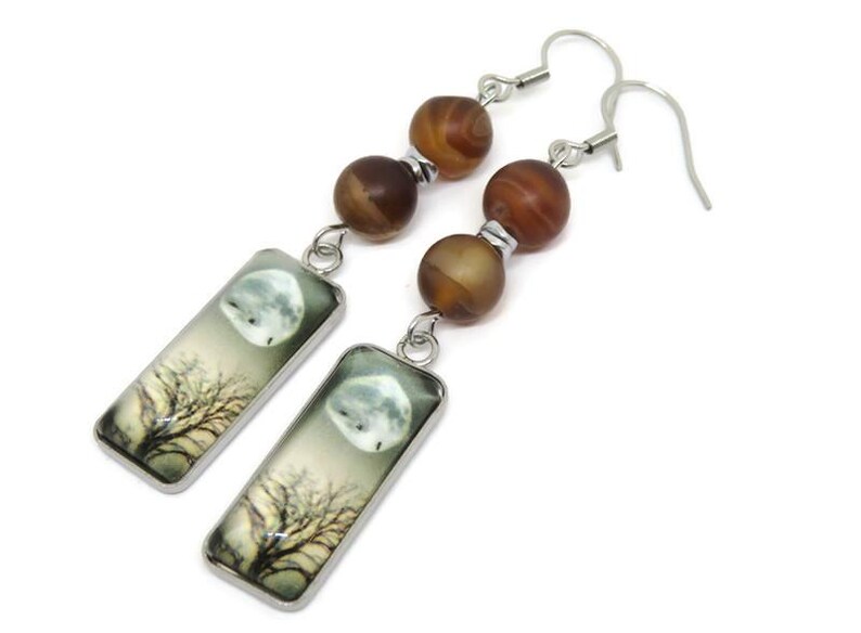 May include: Silver dangle earrings with brown agate beads and rectangular glass charms featuring a moon and tree illustration.