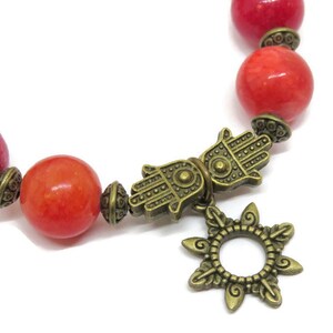 Pu&ograve; includere: Un braccialetto di perline con grandi perline rosse rotonde e dettagli in ottone antico. Il braccialetto include un ciondolo a mano di Hamsa e un ciondolo a forma di sole, creando un'estetica boh&eacute;mien. Le perline hanno un diametro di circa 1 cm.