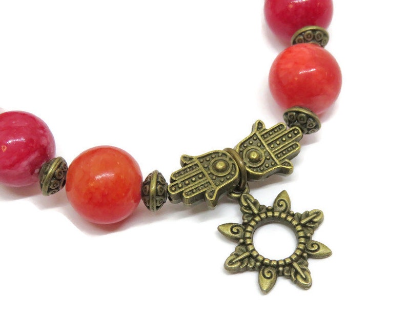 Pu&ograve; includere: Un braccialetto di perline con grandi perline rosse rotonde e dettagli in ottone antico. Il braccialetto include un ciondolo a mano di Hamsa e un ciondolo a forma di sole, creando un'estetica boh&eacute;mien. Le perline hanno un diametro di circa 1 cm.