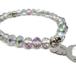 May include: A stretch bracelet composed of faceted, iridescent clear crystal beads with a metallic sheen. The bracelet features a silver-toned metal charm shaped like a hummingbird in flight, attached to the band by a circular metal spacer bead.