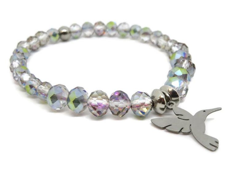 May include: A stretch bracelet composed of faceted, iridescent clear crystal beads with a metallic sheen. The bracelet features a silver-toned metal charm shaped like a hummingbird in flight, attached to the band by a circular metal spacer bead.