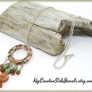 May include: A silver chain necklace with a large, round, brown, pink, and green pendant. The pendant has a hole in the center and is decorated with smaller beads in various colors. The necklace is displayed on a piece of driftwood.