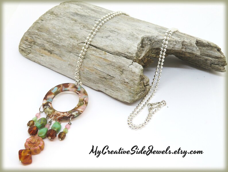 May include: A silver chain necklace with a large, round, brown, pink, and green pendant. The pendant has a hole in the center and is decorated with smaller beads in various colors. The necklace is displayed on a piece of driftwood.