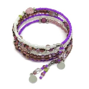May include: A multi-strand wrap bracelet featuring a mix of purple, white, and brown beads. The bracelet has a spiral design with dangling charms and is set against a white background. The beads are of various sizes and textures.