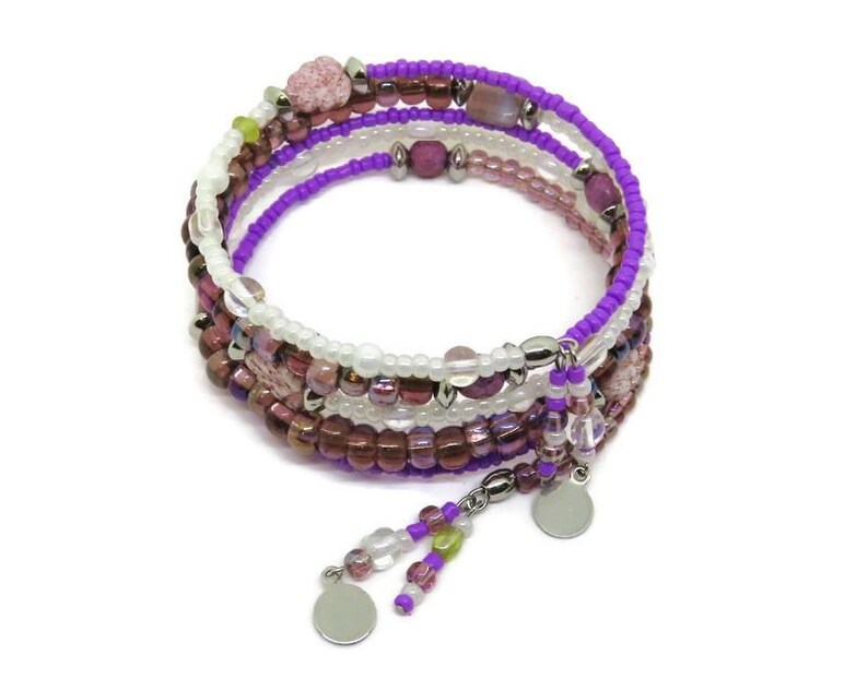 May include: A multi-strand wrap bracelet featuring a mix of purple, white, and brown beads. The bracelet has a spiral design with dangling charms and is set against a white background. The beads are of various sizes and textures.