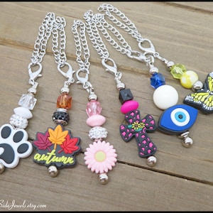 May include: A collection of six silver chain charms with various decorative pendants. Charms include a paw print, autumn leaves, a pink flower, a cross, an evil eye, and a butterfly. The charms are on a wooden surface.