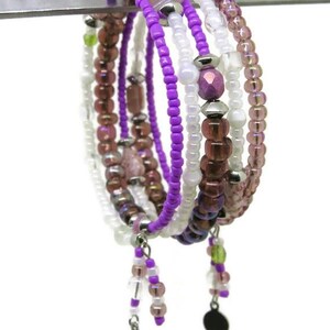 May include: A collection of beaded bracelets in shades of purple, white, and brown. The bracelets feature various bead sizes and shapes, with silver-toned accents and dangling charms. The bracelets are displayed on a silver bar against a white background.