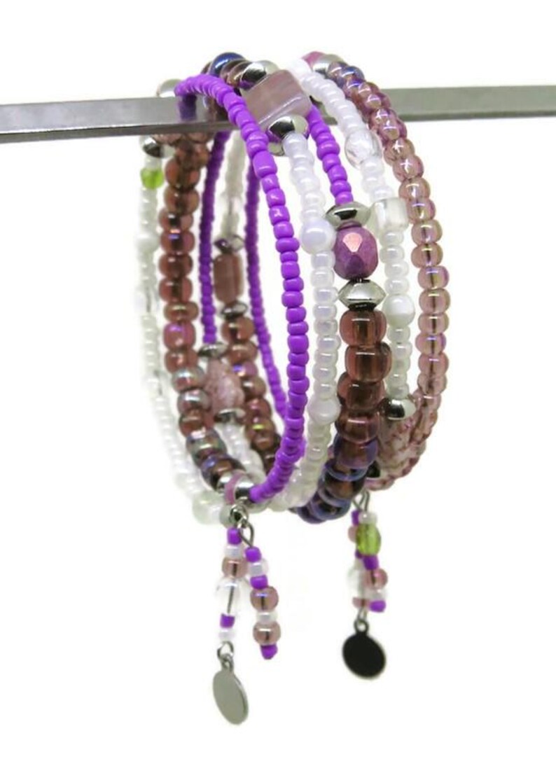 May include: A collection of beaded bracelets in shades of purple, white, and brown. The bracelets feature various bead sizes and shapes, with silver-toned accents and dangling charms. The bracelets are displayed on a silver bar against a white background.