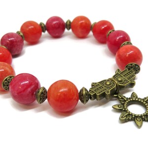 Pu&ograve; includere: Bracciale con perline rotonde rosse e arancioni. Include distanziatori color ottone antico e un ciondolo con una mano di Hamsa e un motivo a forma di sole. Il bracciale &egrave; presentato su sfondo bianco. Le perline misurano circa 1 cm di diametro.