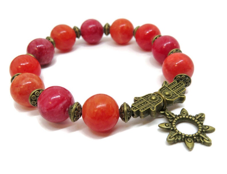 Pu&ograve; includere: Bracciale con perline rotonde rosse e arancioni. Include distanziatori color ottone antico e un ciondolo con una mano di Hamsa e un motivo a forma di sole. Il bracciale &egrave; presentato su sfondo bianco. Le perline misurano circa 1 cm di diametro.