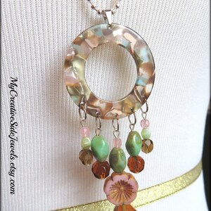 May include: A silver chain necklace with a large round pendant made of clear resin with brown, green, and pink swirls. The pendant has a hole in the center and is attached to a silver chain with a silver clasp. The pendant is decorated with a cluster of green, brown, and pink beads.