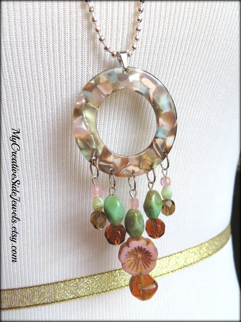 May include: A silver chain necklace with a large round pendant made of clear resin with brown, green, and pink swirls. The pendant has a hole in the center and is attached to a silver chain with a silver clasp. The pendant is decorated with a cluster of green, brown, and pink beads.