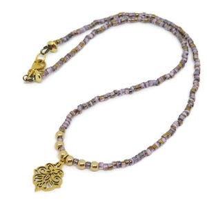 May include: A delicate necklace featuring a gold-colored pendant with an ornate floral design. The necklace is strung with small, iridescent beads in shades of purple, gold, and brown. The clasp is gold-colored.