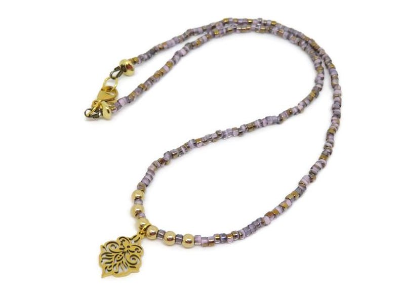 May include: A delicate necklace featuring a gold-colored pendant with an ornate floral design. The necklace is strung with small, iridescent beads in shades of purple, gold, and brown. The clasp is gold-colored.