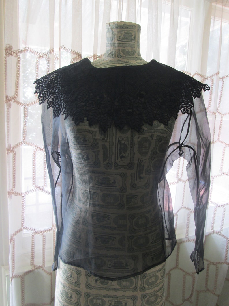 May include: A sheer black blouse with a large, black lace collar. The blouse has long sleeves and is made of a lightweight fabric.